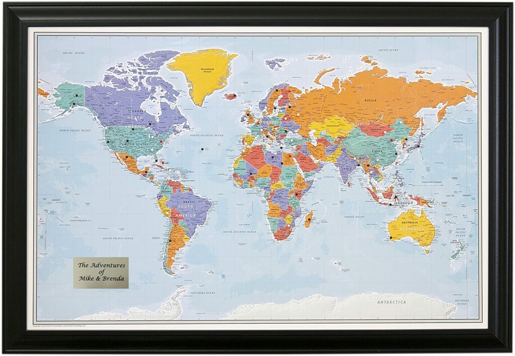 A framed world map with colored countries hanging on a wall, with a push pin inserted into the map.