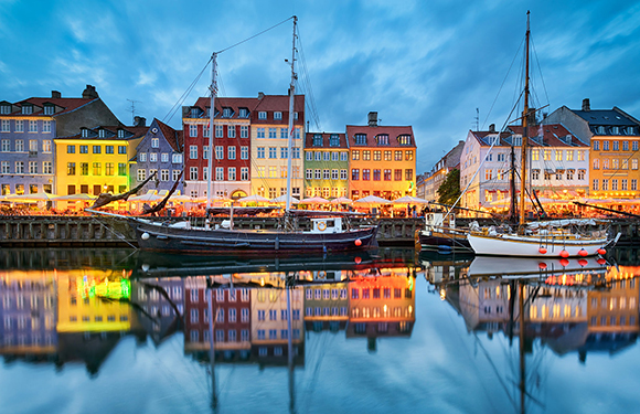 A picturesque view of Copenhagen's colorful buildings reflected in the calm water with boats docked along the quay during twilight.