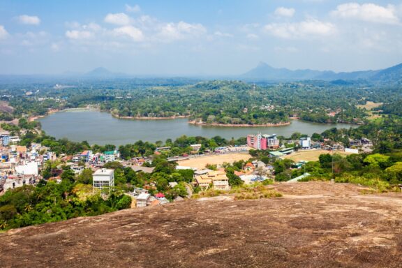 Aerial view of a tropical landscape with a town beside a lake, surrounded by greenery and hills under a blue sky with scattered clouds.