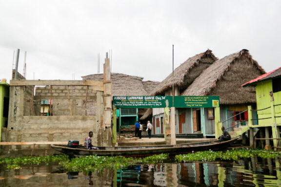 An image of a stilt village with traditional houses and a boat on water covered with green vegetation, under a cloudy sky.