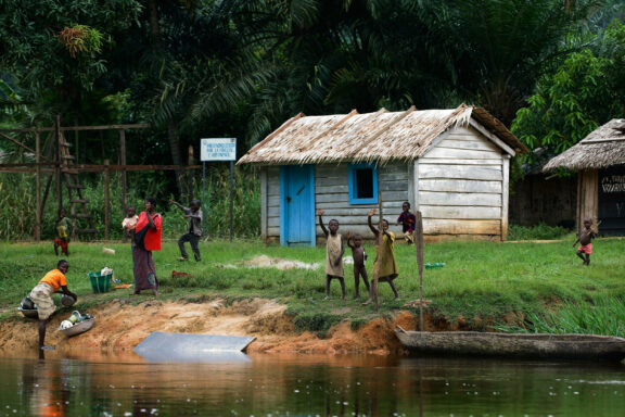 A rural setting with a thatched house by water, people including children outside, some fetching water, surrounded by greenery.