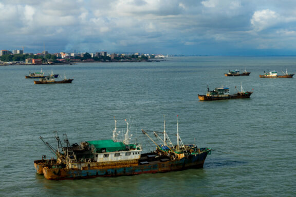 A coastal view featuring several boats on the water with a city skyline in the background under a cloudy sky.