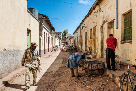 A street scene in Eritrea with people engaged in various activities, buildings lining the street, and a clear sky above.