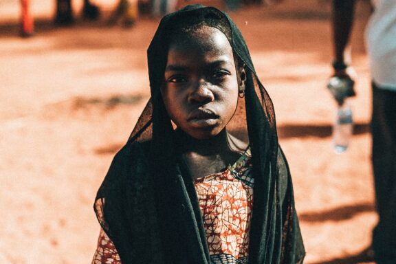 A young person with a headscarf standing outdoors with a slightly blurred background.