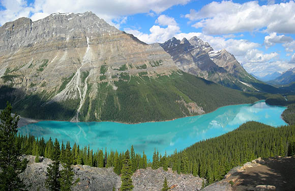 A scenic view of a turquoise lake surrounded by forested mountains under a partly cloudy sky.