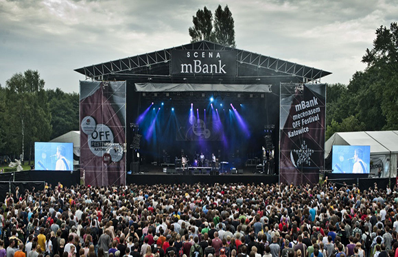 A crowd of people at an outdoor music festival with a stage in the background, large screens on either side, and trees surrounding the area.