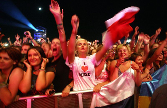 Excited concert crowd cheers, individual in white "I love Muse" shirt waves red cloth.