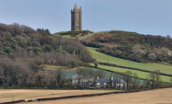 A landscape featuring a tower on a hill with greenery, a few buildings at the hill's base, and a field in the foreground.