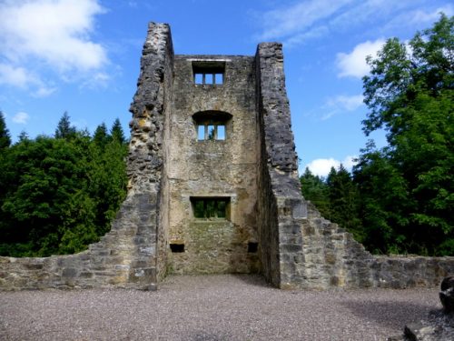 A ruined stone tower with arched windows, surrounded by greenery under a blue sky with clouds.