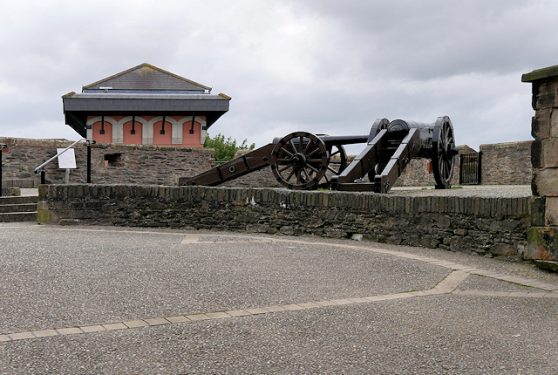 A historical cannon on a carriage is displayed outdoors, against a stone wall and a building with red shutters.