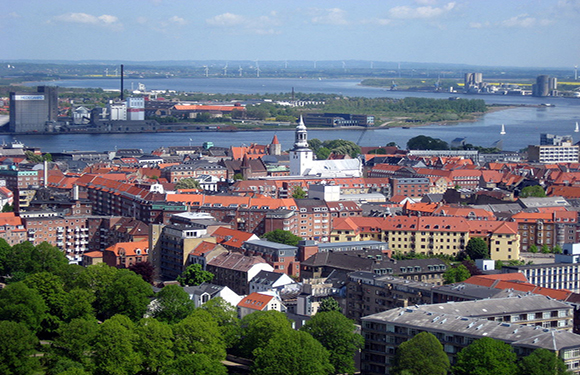 Aerial view of a city with dense building clusters, a prominent church spire, and a large body of water in the background.