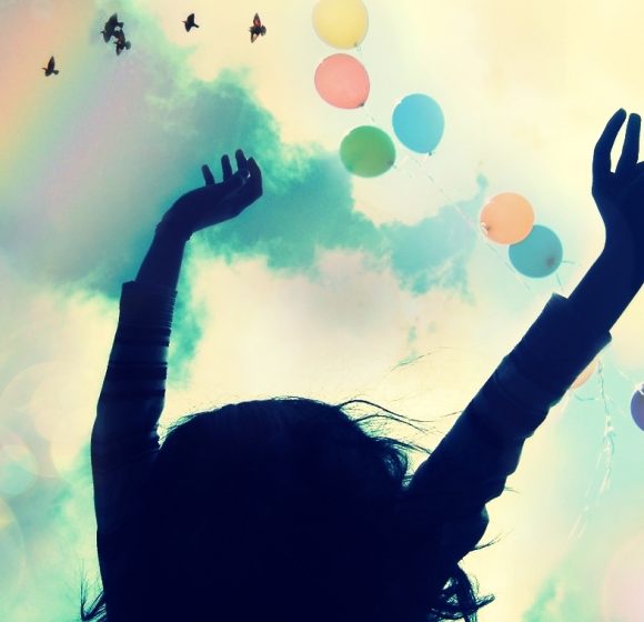 A silhouette of a person with arms raised against a sky with clouds, colorful balloons, and birds flying.