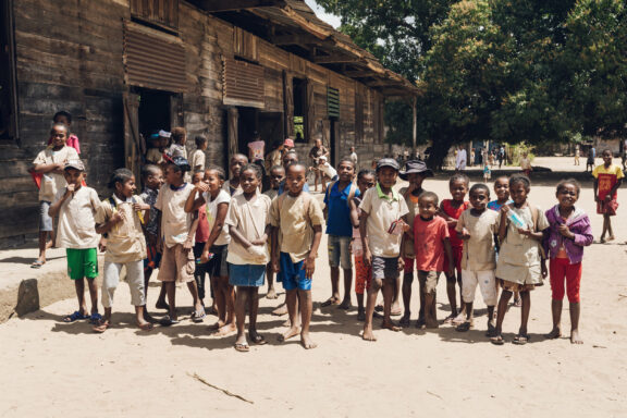 A group of children standing in front of a building with wooden walls and a group of people in the background.