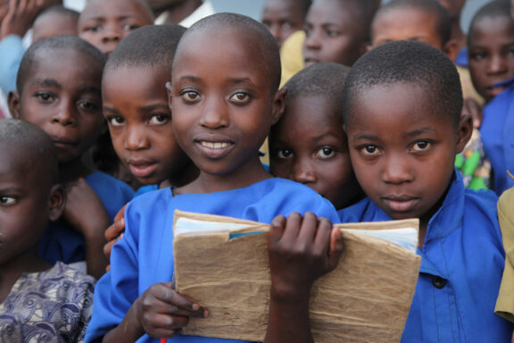 A group of young children with dark hair, some holding books, wearing blue and navy clothing, looking at the camera with a crowd in the background.