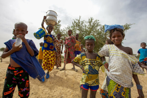 A group of children in colorful clothing, with some carrying items on their heads, walking on sandy ground with sparse vegetation in the background.