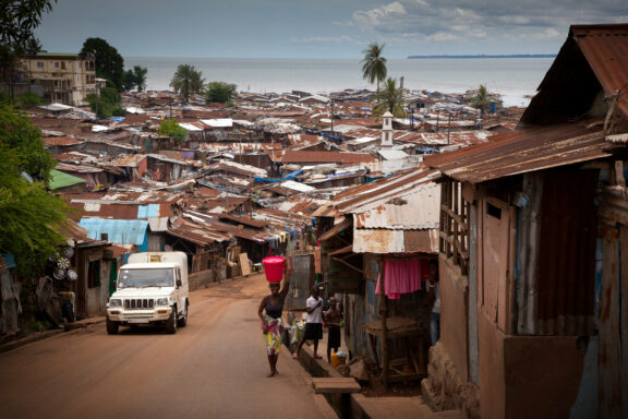 A person walks with a load on their head in a crowded Sierra Leone area with metal roofs, a vehicle is on the left.