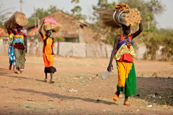 Three individuals walking along a dirt path, each carrying items on their heads, with trees and a clear sky in the background.