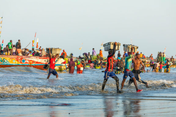 A bustling coastal scene with people on a beach and colorful boats, some walking in water and others on boats, possibly fishing.