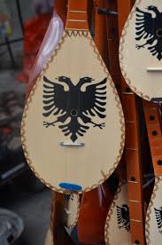 A traditional Albanian çifteli, a stringed instrument with a double-headed eagle design, is set against a blurred outdoor backdrop.