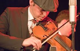 A man in a suit and flat cap playing the violin with focus and intensity.