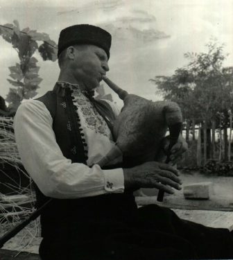 A black and white photo of a man sitting outdoors wearing a traditional vest and cap, playing a bagpipe.