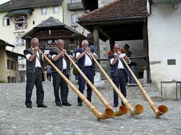 Four individuals playing long wooden alphorns in a traditional outdoor setting with buildings in the background.