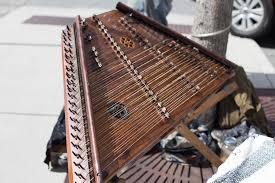 A damaged hammered dulcimer lying on a metal bench outdoors, with some parts broken and strings detached, suggesting abandonment or neglect.