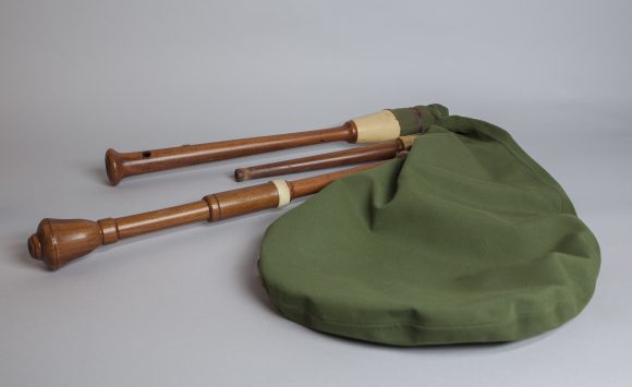 A green fabric bag with two wooden wind instruments, likely parts of a bagpipe, on a grey background.