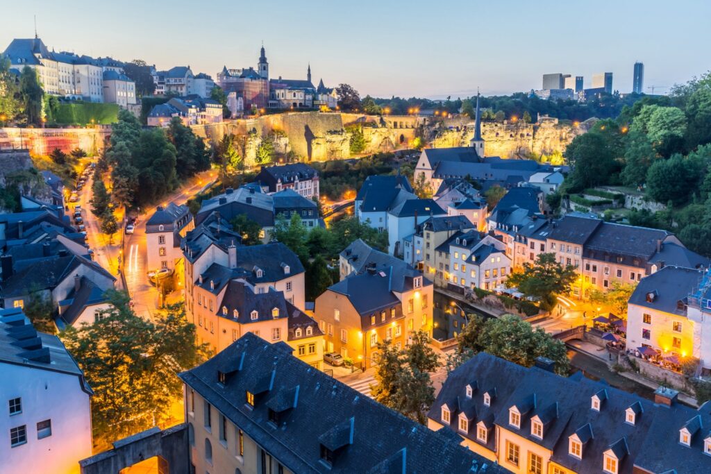 A twilight view of Luxembourg city highlights its historic architecture, illuminated buildings, street lights, and lush greenery.