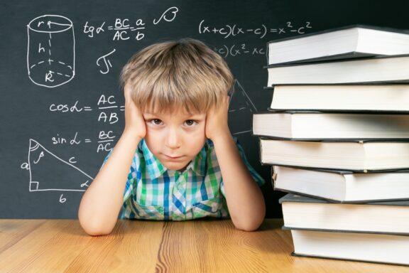 A frustrated boy sits at a desk by a chalkboard full of math equations, with a stack of books beside him.