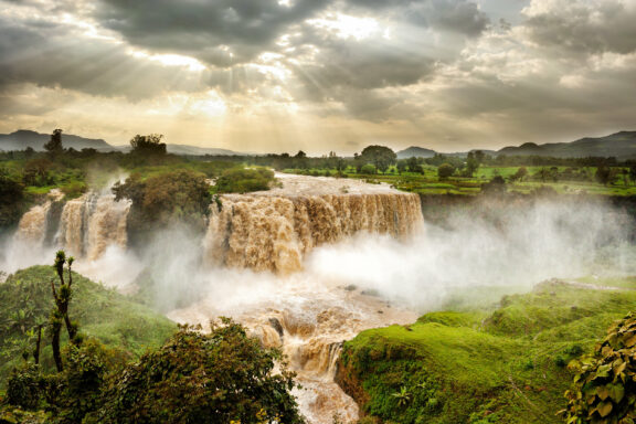 Sunlight pierces clouds above the misty Blue Nile Falls near Bahir Dar, surrounded by lush greenery.