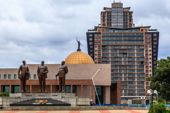 A photo of three statues in front of a building with a golden dome and a tall modern building in the background, located in Gaborone, Botswana.