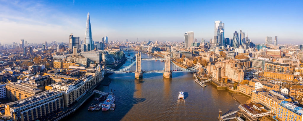 A panoramic aerial view of London showcasing the River Thames, Tower Bridge, and the Shard with clear skies.