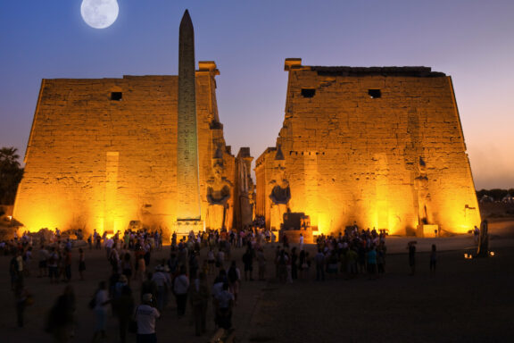 People gathered at dusk at the lit Luxor Temple in Egypt, under a full moon, with an obelisk and statues visible.