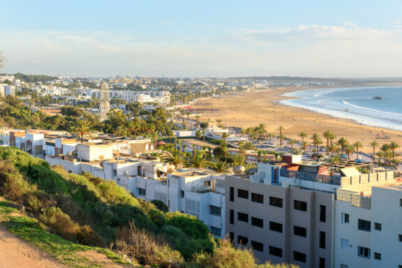 A coastal view of Agadir, Morocco, featuring a beach, buildings, and vegetation under a clear sky.