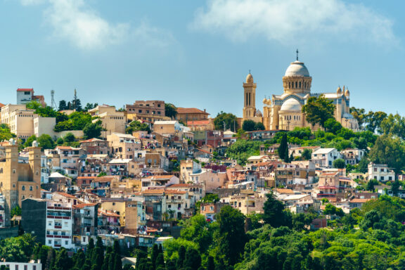 A scenic view of Algiers, Algeria, showcasing densely packed buildings on a hill with a prominent cathedral and minaret under a clear blue sky.