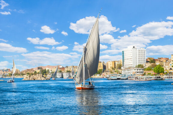 A traditional felucca sailboat on the Nile River with Aswan, Egypt's cityscape in the background.