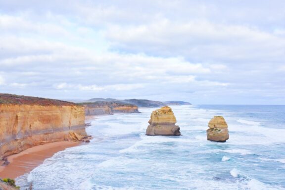 A scenic view of a rugged coastline in Australia, featuring two large rock formations in the ocean near the shore under a cloudy sky.