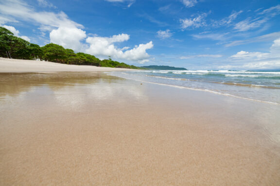 A peaceful beach with reflective wet sand, gentle waves, lush trees, under a partly cloudy sky, possibly in Santa Teresa.