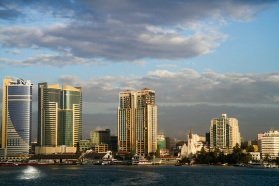 A skyline view of Dar es Salaam with modern high-rise buildings near the waterfront under a partly cloudy sky.