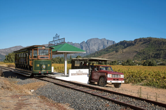 A vintage-style tram is parked at a rural "Franschhoek" station, with a red truck nearby. The backdrop features a vineyard and mountains.