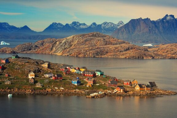 A scenic view of a quaint Greenland coastal village with vibrant houses, calm waters, and mountain backdrop.