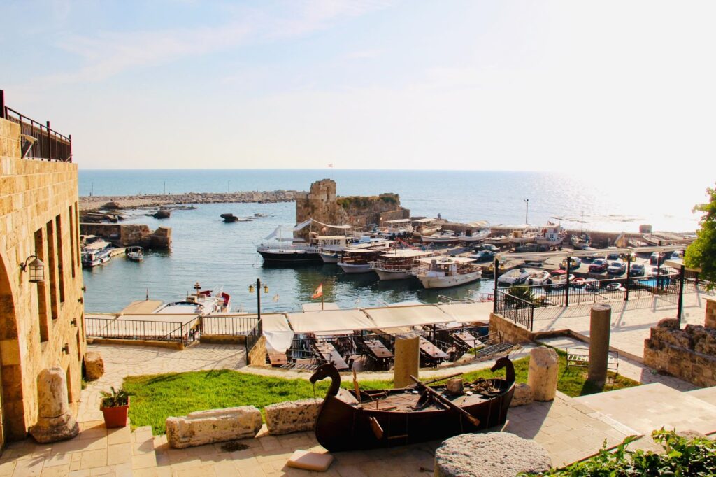 View from a high point of a coastal scene with docked boats, a stone building, and ancient ruins under a clear sky.