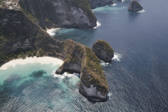 Aerial view of a rugged coastline in Indonesia, featuring a prominent cliff and a secluded beach with turquoise waters.