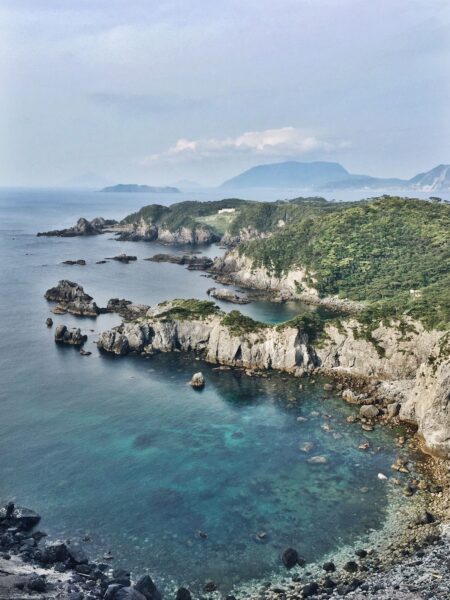 A scenic view of a rocky shoreline in Japan with clear turquoise waters, lush greenery, and mountains in the distance under a hazy sky.