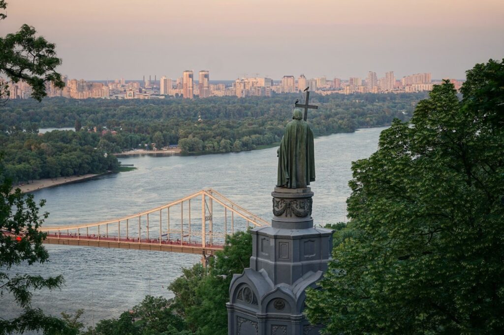 A dusk view of the Dnieper River, a distant bridge, a statue, trees, and Kyiv's cityscape on the horizon.