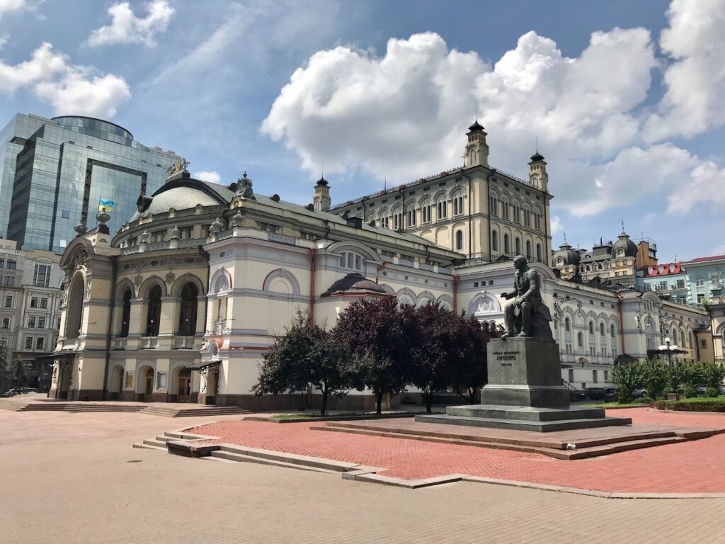 A photo showcases Ukraine's National Opera and Ballet Theater's ornate architecture, a statue, modern buildings, and a cloudy blue sky.