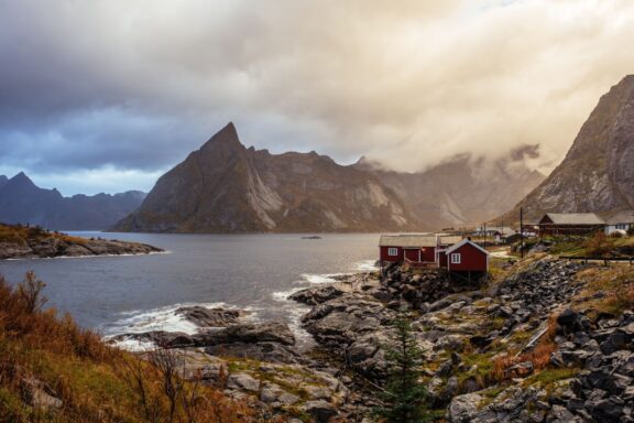 A picturesque Norwegian coastline with rugged cliffs, calm sea, red houses, and a dramatic sky with sun rays through clouds.