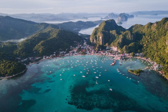 Aerial view of a tropical sea with boats near the coast, surrounded by lush green hills, likely in the Philippines.