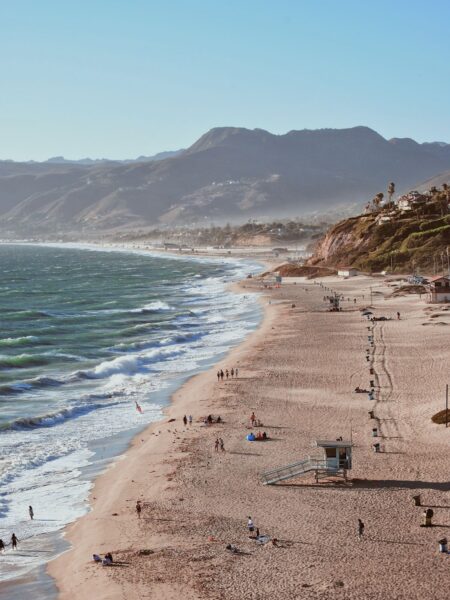 A beach in the USA with sandy shore, waves, and distant mountains, under a clear sky with a few people and beach accessories scattered around.